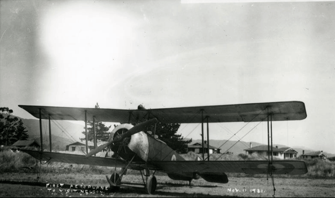 Black and white photo a biplane parked in a field surrounded by a crowd.